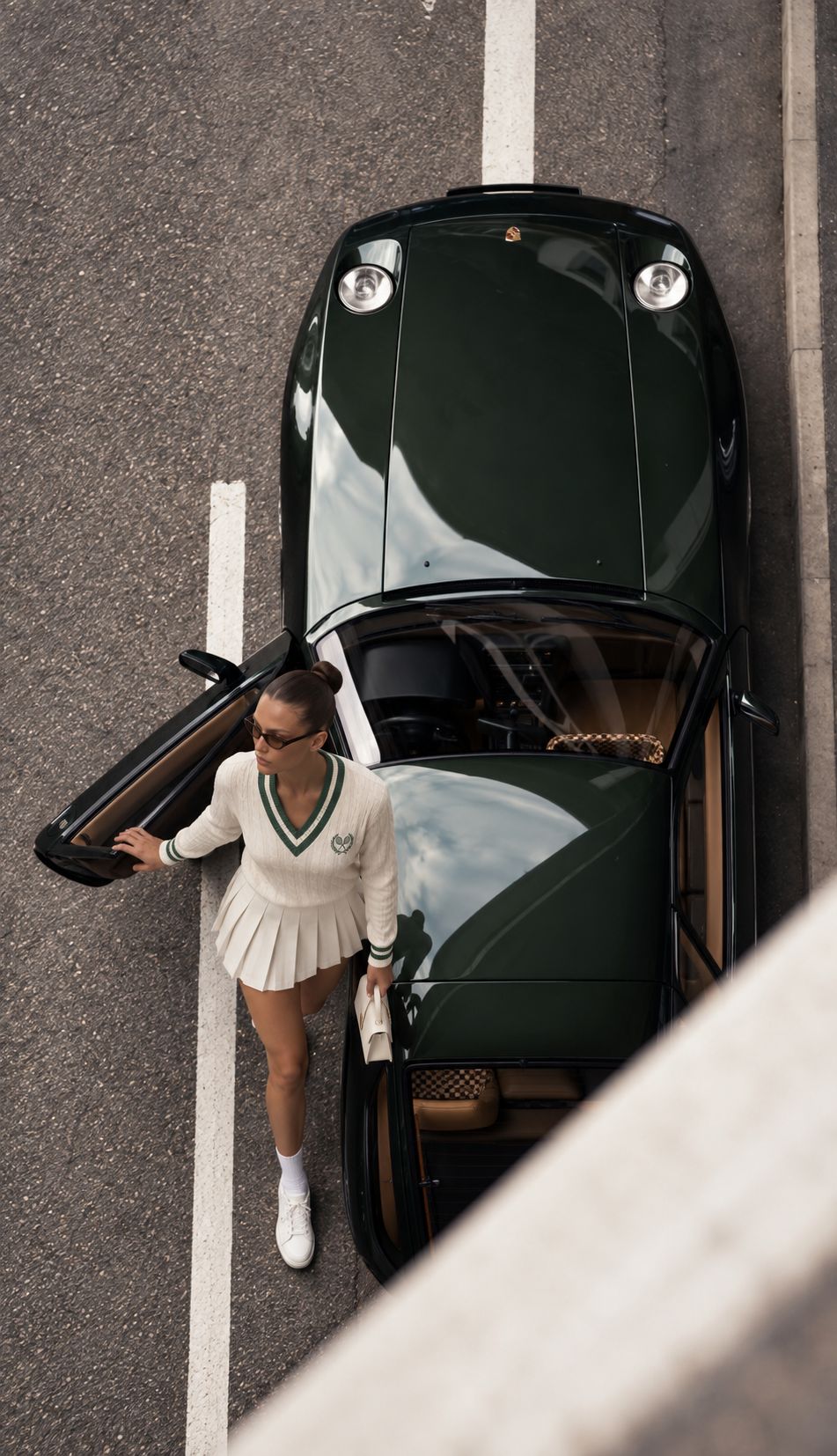 A woman in a cream tennis sweater stepping out of a dark green vintage sports car