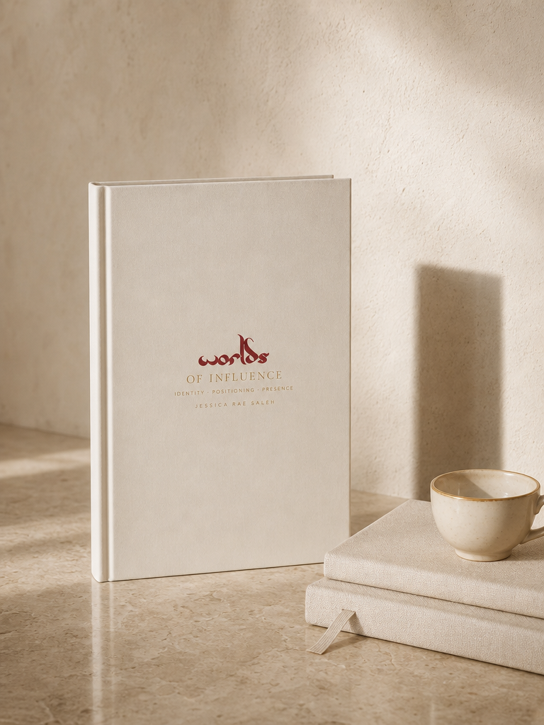 The Worlds of Influence book, cream cover with oxblood red worlds and gold lettering, standing on travertine beside linen notebooks and a gold-rimmed espresso cup